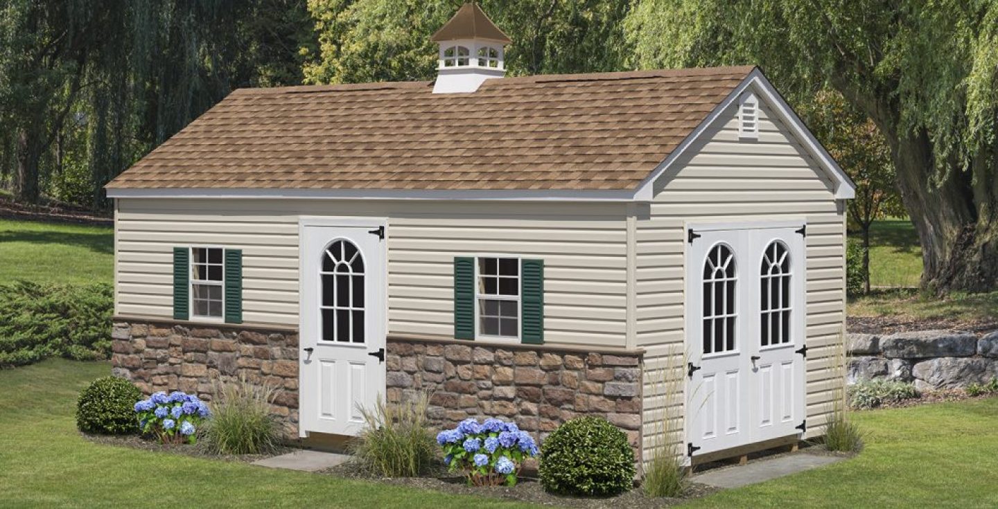 A beige shed with a brown shingle roof, green shutters, white arched doors, stone veneer base, cupola on top, and surrounded by green lawn.