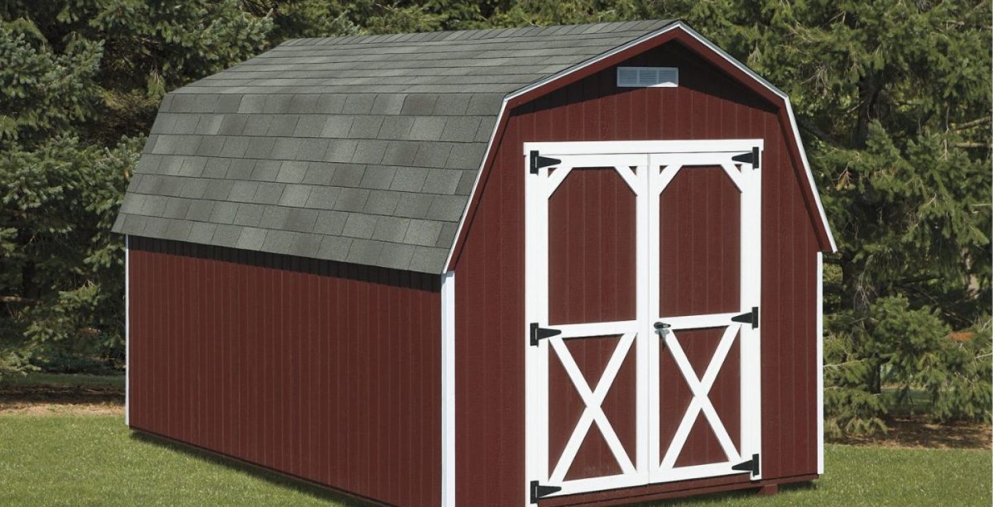 A red shed with a gray gambrel roof, white trim, and double doors featuring white cross-bracing. It is set on a green lawn with trees in the background.