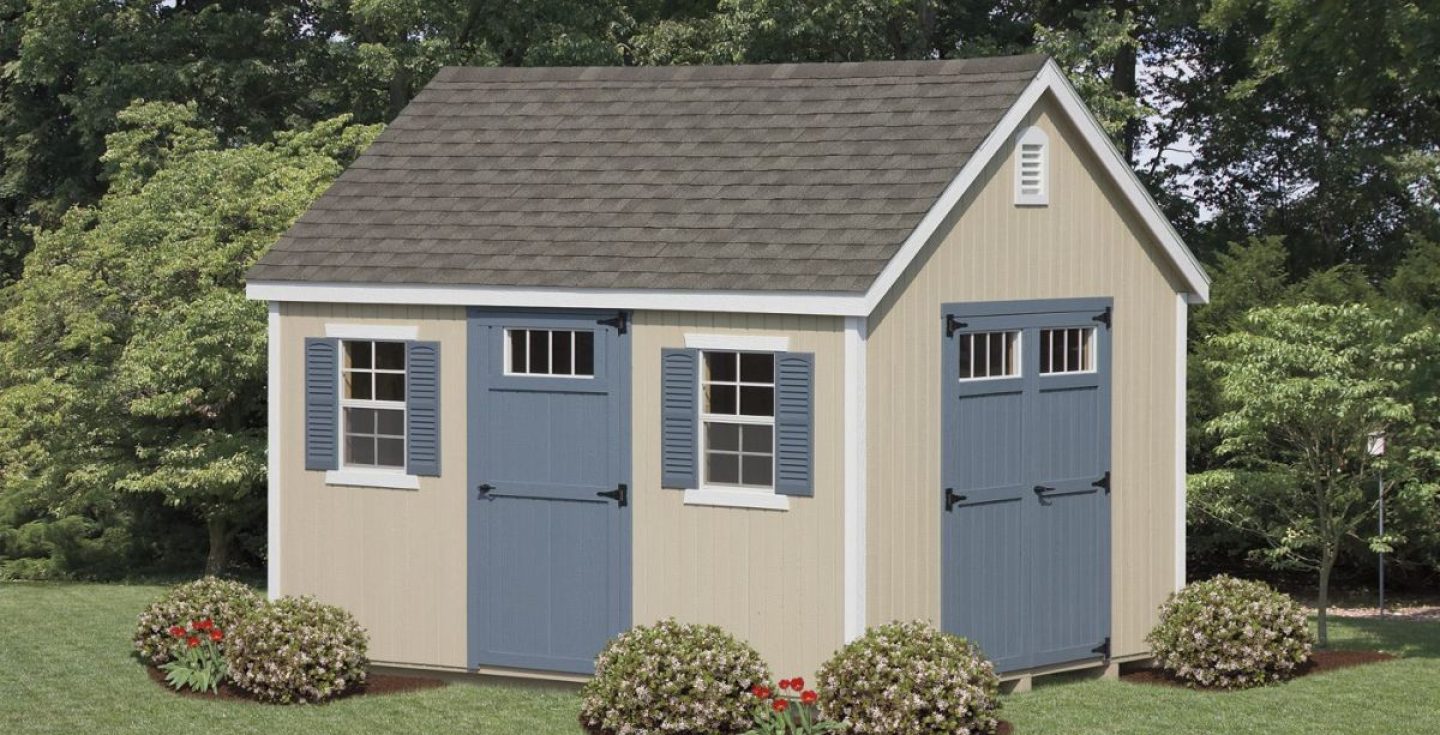 A beige shed with a gray shingle roof, blue shutters, and blue double doors with transom windows. Surrounded by green lawn, bushes, and red flowers.