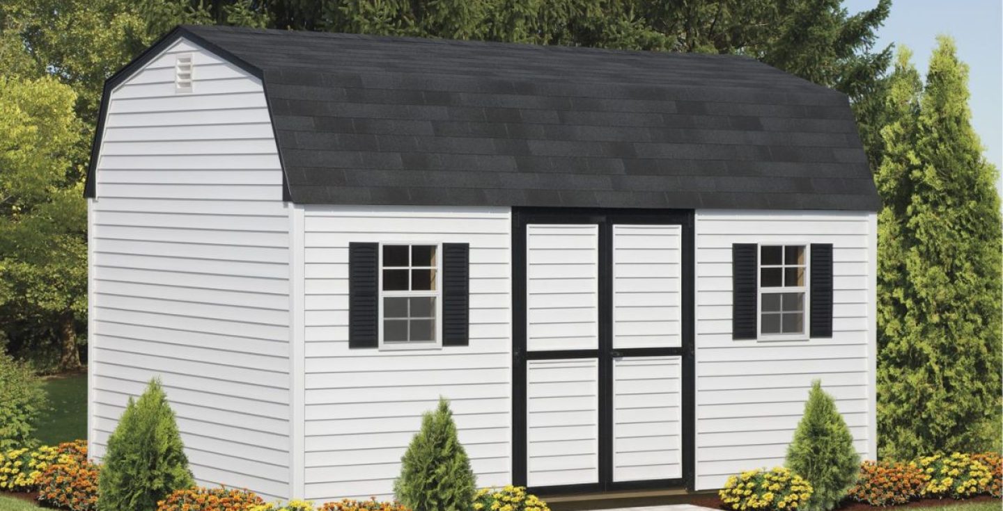 A white shed with a black gambrel roof, black shutters, black double doors, and two windows. It is surrounded by green lawn, shrubs, and yellow flowers.