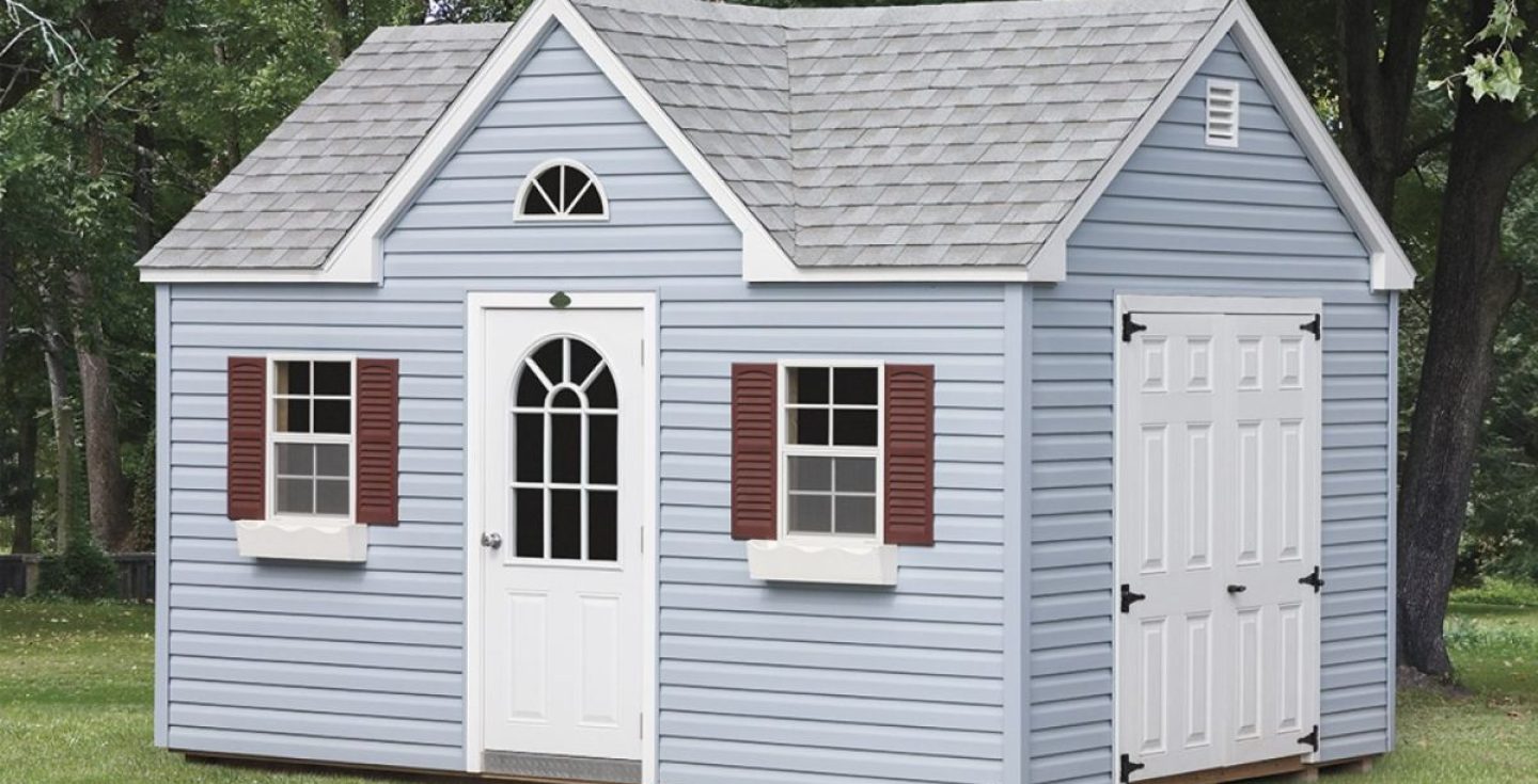 A light blue shed with a gray shingle roof, white arched door, maroon shutters, white trim, and an attic window. It is surrounded by a green lawn and trees.